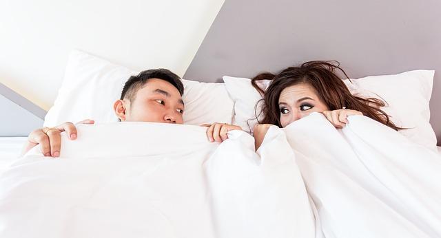 Couple in bed, resting their heads on pillows looking at each other with the bottom part of the face covered by the duvet